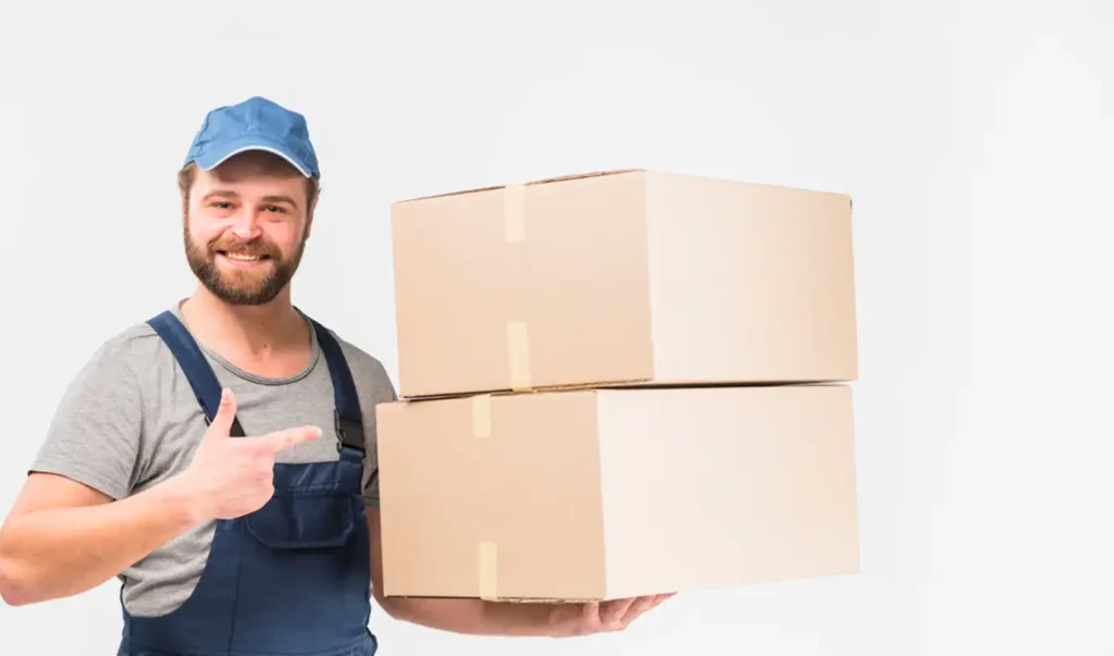 Un livreur souriant, vêtu d'une casquette bleue et d'une salopette, tient deux cartons empilés. Il pointe les cartons du doigt, affichant une attitude amicale et serviable.
